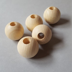 Op de afbeelding: Vijf onafgewerkte houten bollen met een natuurlijke houtnerf. Elke bol heeft een gat in het midden. De bollen zijn gerangschikt op een grijs oppervlak. Deze houten bollen zijn ideaal voor knutselprojecten.