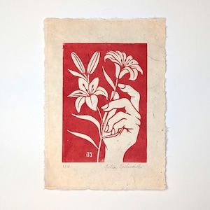 Puede incluir: Un grabado en linóleo rojo y blanco que representa una mano sosteniendo lirios. La impresión está sobre papel texturizado con bordes irregulares. La firma del artista y el número de edición son visibles.