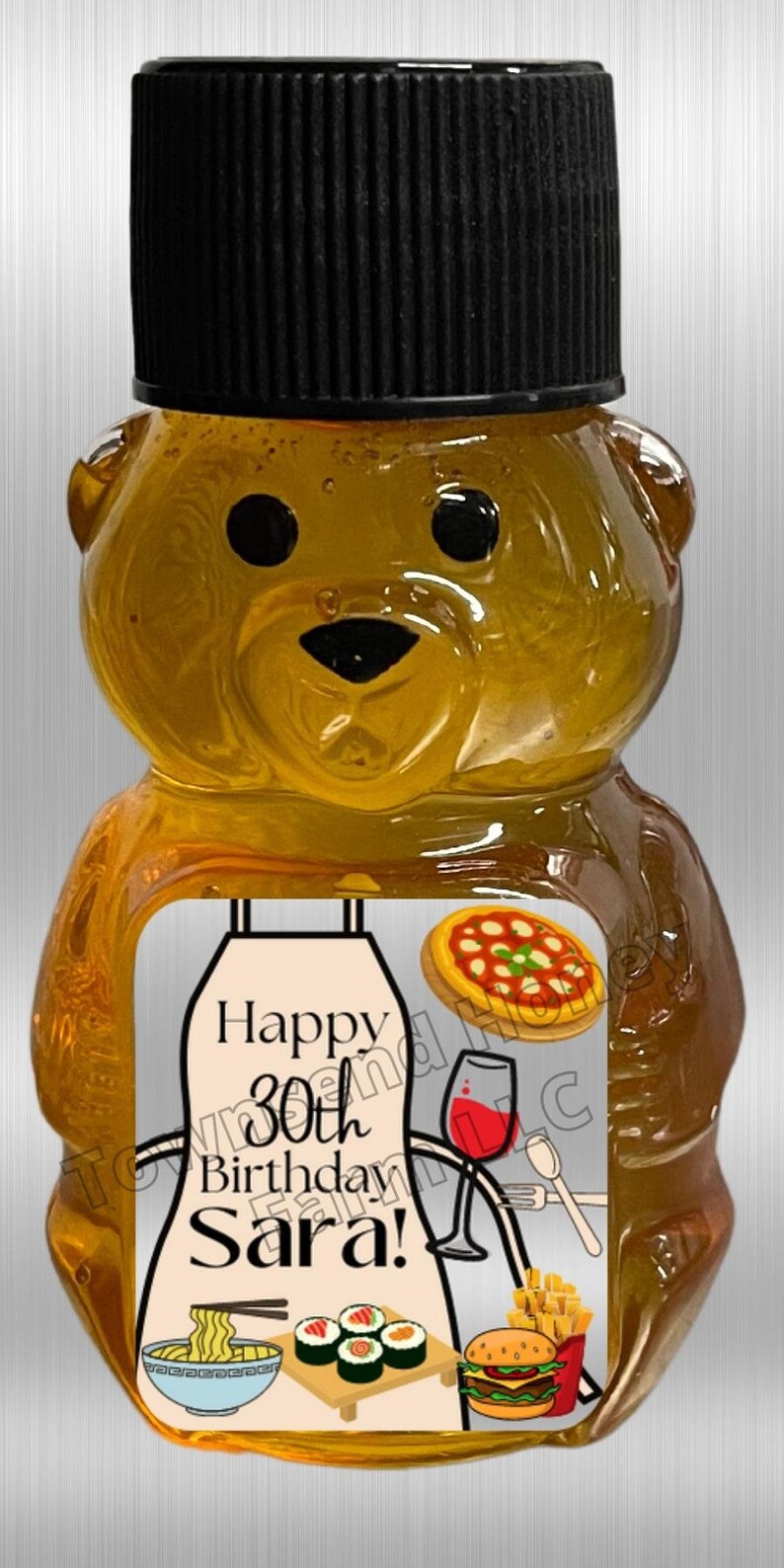 May include: A honey bear-shaped bottle with a black cap. The label reads "Happy 30th Birthday Sara!" and features illustrations of food items like pizza, sushi, a burger, and noodles. The bottle is filled with golden honey.