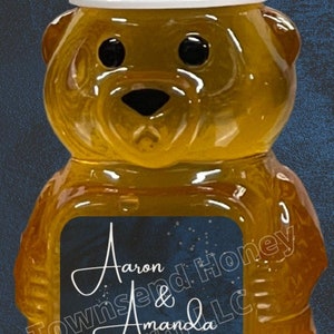 May include: A bear-shaped honey bottle with a white cap. The bottle has a label with the names "Aaron & Amanda Forseth" and the date "7.3.2022". The honey is a golden color.