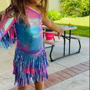 May include: A young girl wearing a one-shoulder iridescent blue and pink top with a fringe skirt. She is wearing pink shoes with a gold bow.