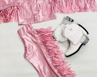 Puede incluir: Un conjunto de vaquera de satén rosa con detalles de flecos. El conjunto incluye una chaqueta, pantalones y un sombrero de vaquero blanco con una estrella plateada.