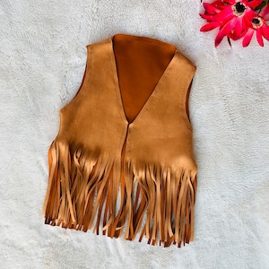Peut inclure: Un gilet en daim marron avec des détails en franges. Le gilet a un col en V et est sans manches.