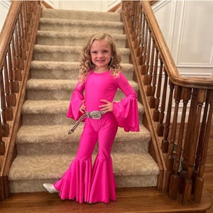 May include: A young girl wearing a hot pink jumpsuit with flared trousers and long sleeves. She is wearing a silver sequined belt and has her hand on her hip. She is standing on a carpeted staircase.