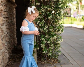 May include: A young girl wearing a light blue denim overall dress with a white lace trim on the shoulders. The dress has a flared skirt and the girl is wearing a white bow in her hair.