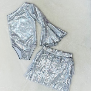May include: A silver sequined bodysuit with a single long bell sleeve and a matching silver sequined skirt with fringe.