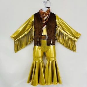 Puede incluir: Un mono dorado con flecos en las mangas y pantalones acampanados. El mono tiene un chaleco de ante marrón con flecos en la parte inferior. Una bufanda marrón estampada está atada alrededor del cuello.