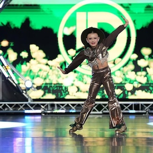 Könnte beinhalten: Eine junge Person in einem braunen Cowboyhut und einem braunen und silbernen Outfit tanzt auf einer Bühne. Die Bühne hat einen grünen Bildschirm hinter sich mit einem großen weißen Kreis mit einem schwarzen Buchstaben D in der Mitte.