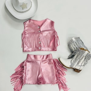 Puede incluir: Conjunto de chaleco y pantalones cortos de imitación de cuero rosa con ribete de flecos. El chaleco tiene cierre de botón y los pantalones cortos tienen cintura alta.