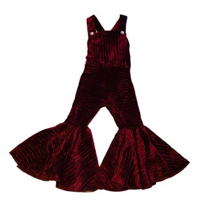 May include: Burgundy velvet bell bottom overalls with a wide leg and a ruffled hem. The overalls have a simple design with two straps and two buttons.