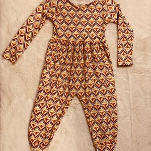May include: A long-sleeved jumpsuit for a child, featuring a geometric pattern in shades of brown, yellow, and white. The jumpsuit has a round neckline and a gathered waist.