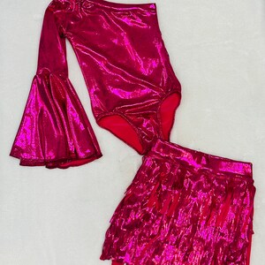May include: A pink one-shoulder bodysuit with a long bell sleeve and a matching pink fringed skirt.