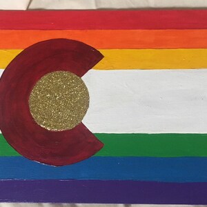 Op de afbeelding: Een handgeschilderd houten bord met de vlag van de staat Colorado met een regenboog-pride-vlagontwerp. De vlag is geschilderd op een houten plank met een streepjespatroon in rood, oranje, geel, groen, blauw en paars. De vlag van de staat Colorado bevindt zich in het midden van het bord met een gouden glittercirkel in het midden van de 'C'.