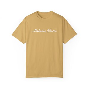 Op de afbeelding: Een mosterdgeel T-shirt met de tekst "Alabama Charm" in witte cursieve letters.