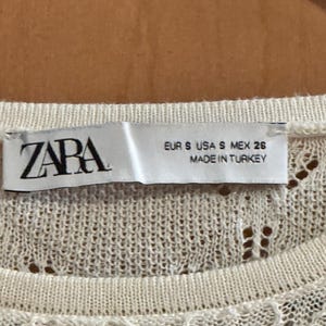 May include: A close-up of a cream-coloured knit top with a delicate openwork pattern. The label reads "ZARA", with size details: EUR S, USA S, MEX 26. The tag also states the item was made in Turkey.