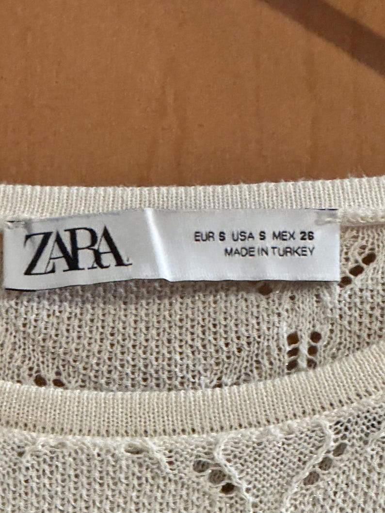 May include: A close-up of a cream-coloured knit top with a delicate openwork pattern. The label reads "ZARA", with size details: EUR S, USA S, MEX 26. The tag also states the item was made in Turkey.