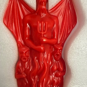 May include: A red devil figurine with a trident, wings, and two smaller figures in flames. The devil has horns and is holding a trident. The figurine is made of a glossy red material.