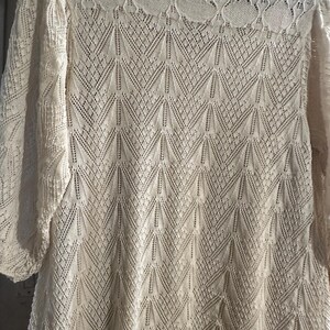May include: A cream-coloured, long-sleeved top with an intricate openwork knit design. The round neckline has a decorative lace pattern. The sleeves are three-quarter length. The top is made of a soft, lightweight material.