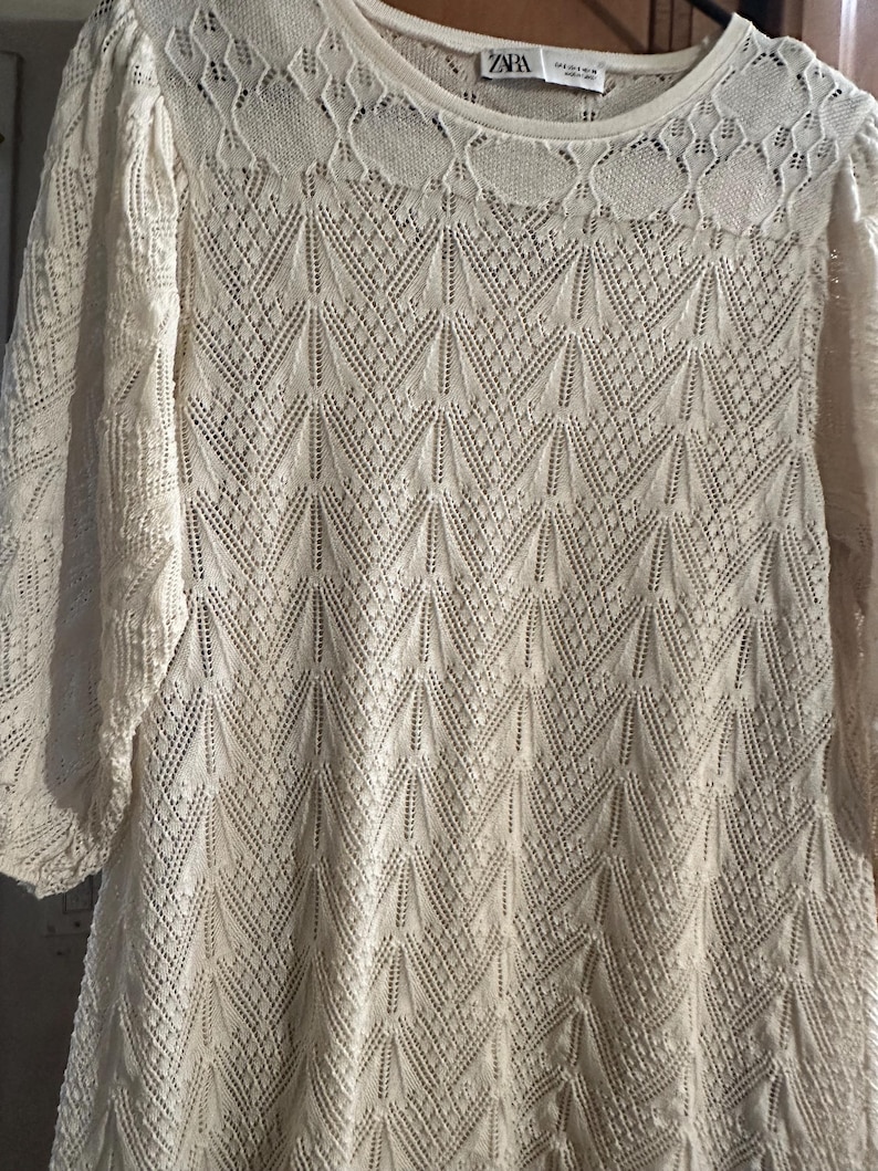 May include: A cream-coloured, long-sleeved top with an intricate openwork knit design. The round neckline has a decorative lace pattern. The sleeves are three-quarter length. The top is made of a soft, lightweight material.