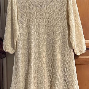 May include: A cream-coloured, knee-length dress with a delicate, open-knit pattern. The dress has three-quarter sleeves and a round neckline. The fabric appears soft and lightweight. The dress is hanging on a hanger.