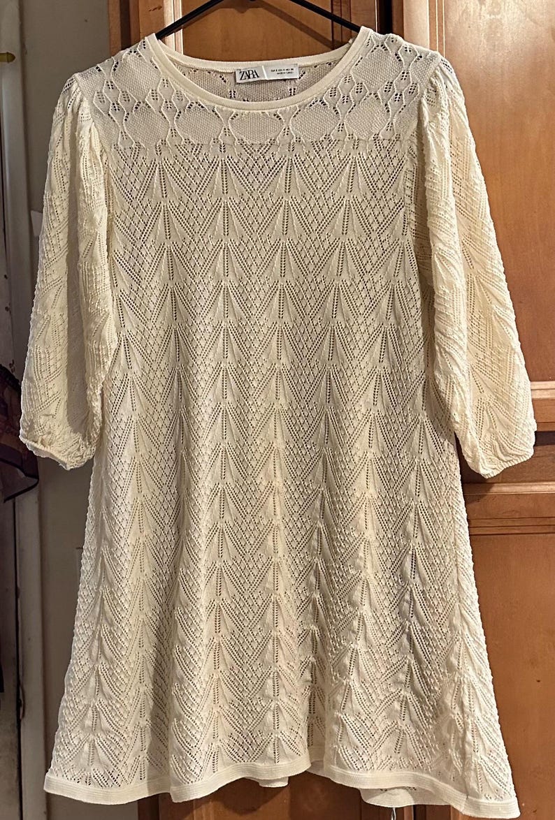 May include: A cream-coloured, knee-length dress with a delicate, open-knit pattern. The dress has three-quarter sleeves and a round neckline. The fabric appears soft and lightweight. The dress is hanging on a hanger.