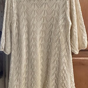 May include: Cream-coloured, long-sleeved dress with a delicate, open-knit pattern. The dress features a round neckline and a slightly flared hem. The sleeves are three-quarter length. The overall design is a geometric pattern.