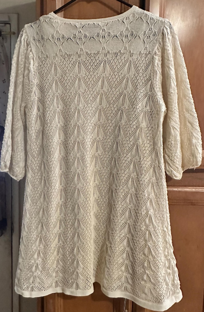 May include: Cream-coloured, long-sleeved dress with a delicate, open-knit pattern. The dress features a round neckline and a slightly flared hem. The sleeves are three-quarter length. The overall design is a geometric pattern.