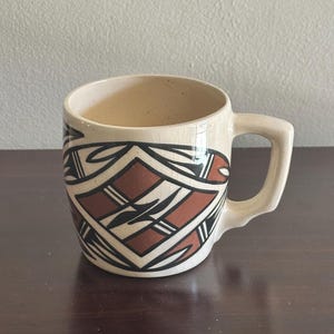 Pode incluir: Caneca de cerâmica creme com um design geométrico em preto e castanho. A caneca tem uma pega e está cheia de um líquido castanho claro. O design apresenta formas de diamante e angulares. A caneca está numa superfície castanha escura.