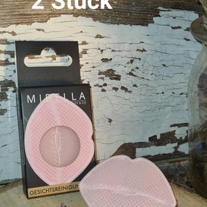 Könnte beinhalten: Zwei rosa Silikon-Gesichtsreinigungswerkzeuge in Lippenform. Die Werkzeuge haben eine strukturierte Oberfläche und sind in einer schwarzen Box mit dem Text "Mibella Beauty & Co" und "Gesichtsreinigungsun" verpackt. Der Text "2 Stück" ist auf einer Holzoberfläche hinter der Box geschrieben.