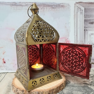 Könnte beinhalten: Eine messingfarbene Metalllaterne mit geometrischen Aussparungen und roten Glasscheiben. Die offene Tür zeigt eine brennende Kerze im Inneren. Die Laterne steht auf einer Holzscheibe und erzeugt eine warme Atmosphäre.