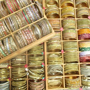 Könnte beinhalten: Eine Holzkiste gefüllt mit Reihen von bunten Armreifen. Die Armreifen sind aus Glas, Metall und Kunststoff und in verschiedenen Farben erhältlich, darunter Gold, Silber, Rot, Grün und Blau.