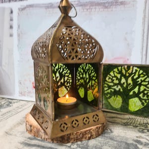 Könnte beinhalten: Eine dekorative Messinglaterne mit einem Baumdesign auf den Glasscheiben. Die Laterne hat eine kleine brennende Kerze im Inneren und steht auf einem Holzsockel. Die Metallarbeiten der Laterne weisen aufwendige Ausschnitte auf.