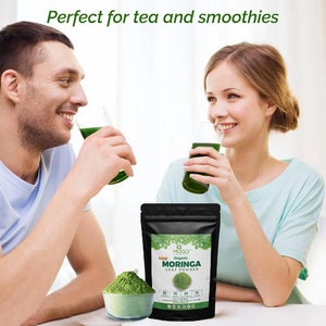 Może przedstawiać: Dwie osoby piją zielone koktajle. Na białym stole przed nimi znajduje się woreczek z MOGO Raw Organic Moringa Leaf Powder. Woreczek jest czarny z zielonym wzorem liścia i nadrukiem "RAW ORGANIC MORINGA LEAF POWDER".