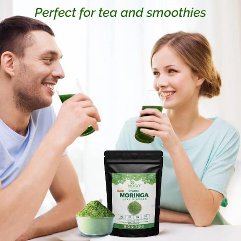 Może przedstawiać: Dwie osoby piją zielone koktajle. Na białym stole przed nimi znajduje się woreczek z MOGO Raw Organic Moringa Leaf Powder. Woreczek jest czarny z zielonym wzorem liścia i nadrukiem "RAW ORGANIC MORINGA LEAF POWDER".