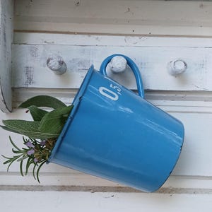 Pode incluir: Uma caneca de metal esmaltado azul brilhante com alça, apresentando o texto branco 0,5 impresso na lateral. A caneca está pendurada num gancho de madeira branca e contém um pequeno ramo de folhas verdes de sálvia e alecrim com flores.