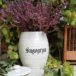 May include: A white ceramic barrel-shaped container with the word "Sagogryn" in black script, filled with purple flowers. Green hops and foliage surround the container, with a white lid nearby.