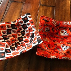 May include: Two square bowl cozies. One features a black and white checkerboard pattern with orange beaver logos. The other is orange with OSU, Oregon State, and beaver graphics. Both are quilted.