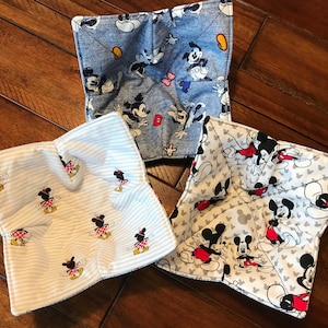 May include: Three square fabric bowl cozies with Mickey and Minnie Mouse patterns. One is denim blue with Mickey Mouse, another is white with gray stripes and Minnie Mouse, and the third is white with Mickey Mouse.