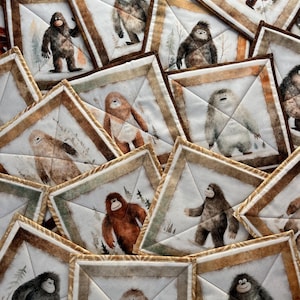 May include: A collection of square fabric coasters featuring illustrations of Bigfoot in various poses. Each coaster has a white background with a forest scene and a brown border. The Bigfoot figures are depicted in shades of brown and gray.