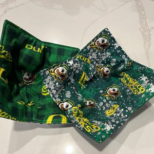 May include: Two square fabric bowl cozies. One is green plaid with yellow text and the word "Ducks". The other is green with white splatters and Oregon Ducks logos. Both are on a white surface.