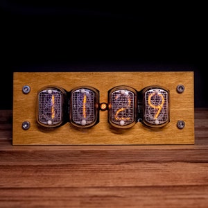 May include: A wooden clock with four Nixie tubes displaying the time 11:29. The clock has a light brown wood finish and is set against a dark background.