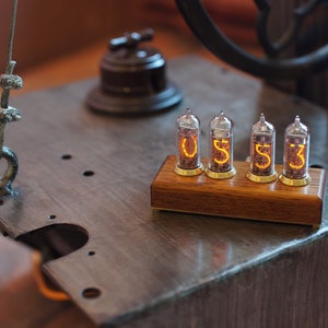 May include: A wooden base with four clear glass Nixie tubes displaying the numbers "0533". The tubes are lit with a warm orange glow.