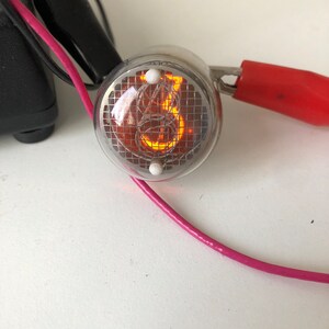 Puede incluir: Primer plano de un tubo Nixie de color plateado que muestra el n&uacute;mero "8". El tubo est&aacute; iluminado con un brillo naranja y tiene un patr&oacute;n de cuadr&iacute;cula visible en su interior. El tubo est&aacute; conectado a un cable rosa y un cable rojo con una abrazadera.