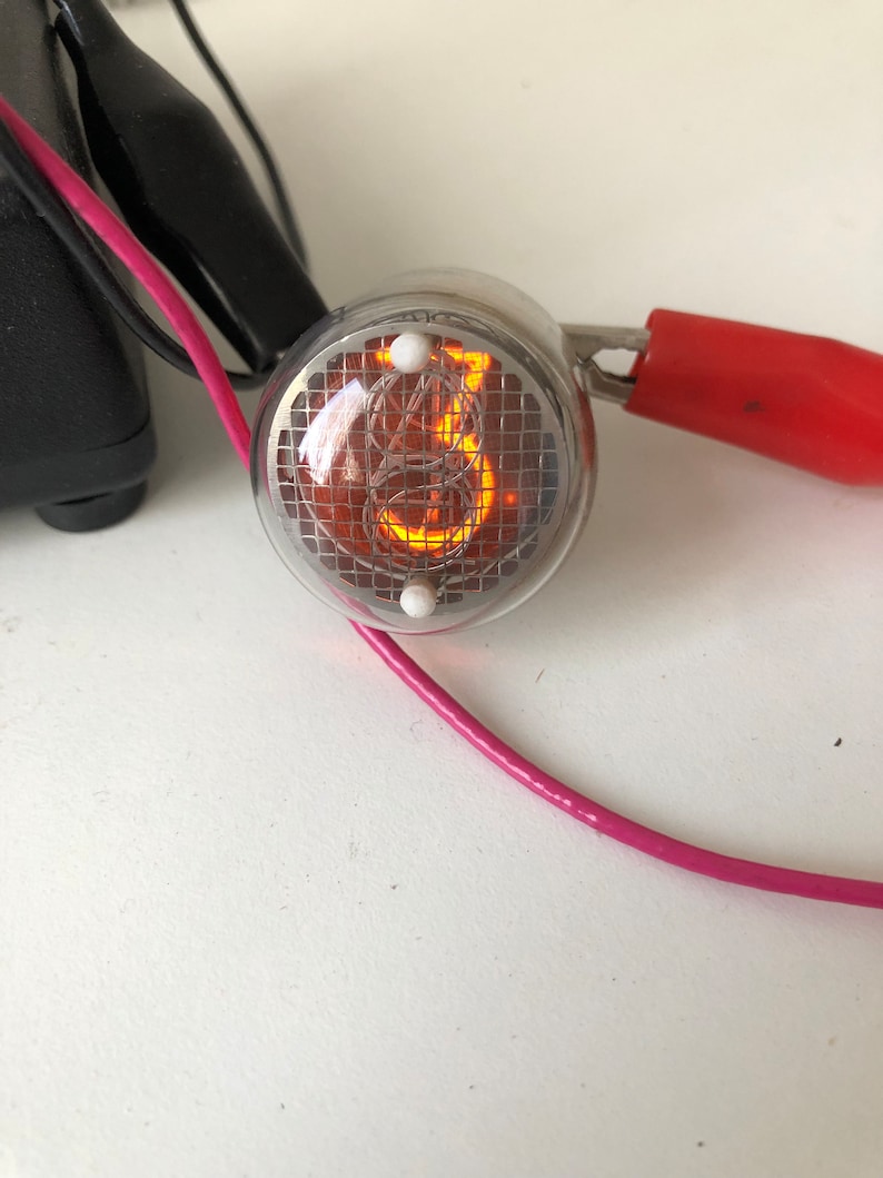 Puede incluir: Primer plano de un tubo Nixie de color plateado que muestra el n&uacute;mero "8". El tubo est&aacute; iluminado con un brillo naranja y tiene un patr&oacute;n de cuadr&iacute;cula visible en su interior. El tubo est&aacute; conectado a un cable rosa y un cable rojo con una abrazadera.