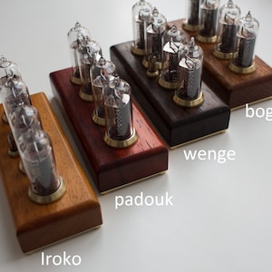 Puede incluir: Cuatro bases de madera para un reloj de tubo Nixie, cada una con un tipo de madera diferente: Iroko, Padouk, Wengu&eacute; y Roble de pantano. Las bases est&aacute;n dispuestas en una fila de izquierda a derecha, con la base de Iroko en la parte inferior y la base de Roble de pantano en la parte superior.
