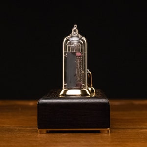Puede incluir: Un reloj de color dorado con un tubo de vidrio transparente que contiene un elemento rojo incandescente. El reloj est&aacute; sentado sobre una base de madera oscura.