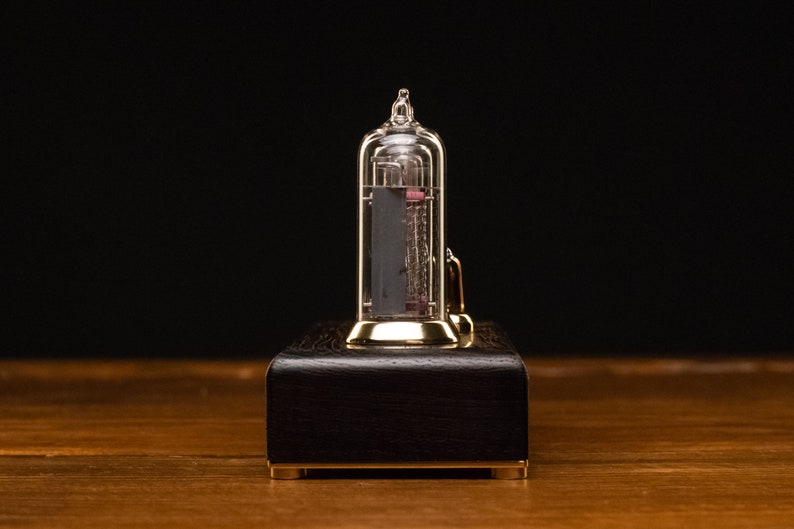 Puede incluir: Un reloj de color dorado con un tubo de vidrio transparente que contiene un elemento rojo incandescente. El reloj est&aacute; sentado sobre una base de madera oscura.