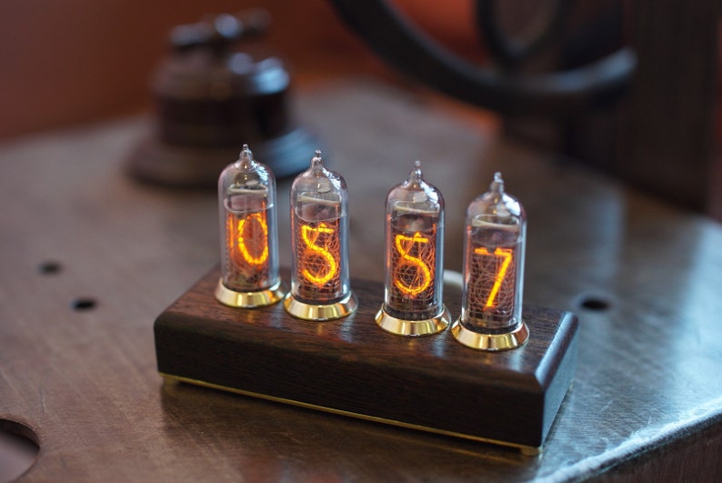 May include: A wooden clock with four Nixie tubes displaying the time 05:57. The tubes are lit with an orange glow and are set in a dark wood base.