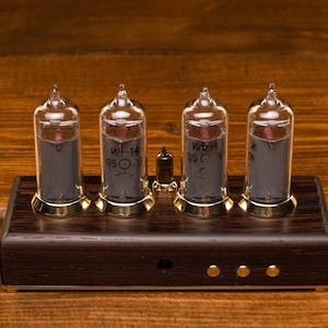 Puede incluir: Un reloj de cuatro d&iacute;gitos con cuatro tubos Nixie de vidrio transparente que muestran la hora. Los tubos est&aacute;n montados en una base de madera oscura con detalles dorados. Los tubos est&aacute;n etiquetados con "ИН-14 05 С".