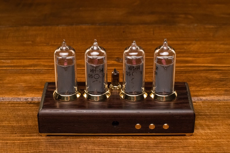 Puede incluir: Un reloj de cuatro d&iacute;gitos con cuatro tubos Nixie de vidrio transparente que muestran la hora. Los tubos est&aacute;n montados en una base de madera oscura con detalles dorados. Los tubos est&aacute;n etiquetados con "ИН-14 05 С".